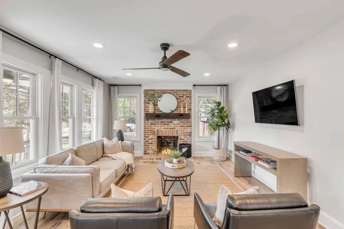 This image features a stylish living room with a brick fireplace, a comfortable sofa, a coffee table, a TV, and a touch of greenery from a potted plant. Sunlight streams in, highlighting the spacious layout and creating a welcoming, relaxed mood. The neutral décor and plush furnishings set a sophisticated, cozy atmosphere perfect for group gatherings.