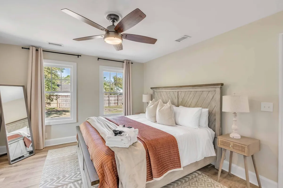 A serene bedroom with plush bedding as the focal point, large windows allowing natural light to flood in, a ceiling fan overhead, and a large mirror that enhances the airy atmosphere. Soft textures and a refined, cozy palette create an inviting environment.