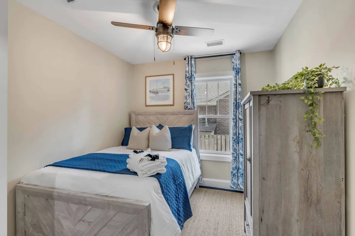 A relaxing bedroom featuring a bed with a blue quilt, a window with soft curtains letting in gentle daylight, a decorative plant, and framed artwork. The overall mood is calm and cozy, ideal for unwinding.