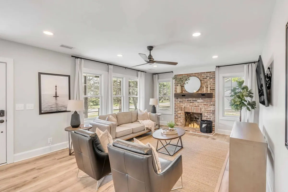 A bright, comfortable living space with a soft sofa, brick fireplace, large windows letting in abundant natural light, a coffee table, and colorful artwork. The open design and elegant décor create a friendly, social ambiance perfect for both lively conversations and quiet relaxation.