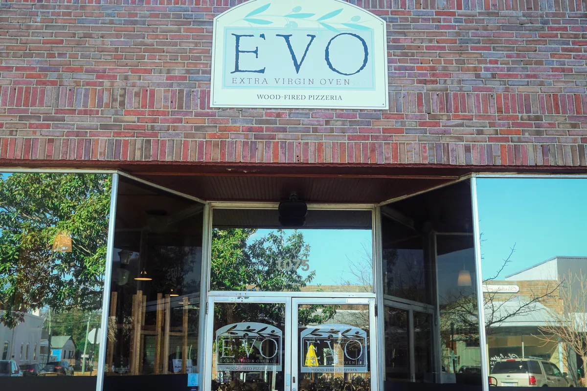 This image displays the welcoming exterior of a restaurant with a visible sign, glass entrance doors, and a brick facade. Diners are seen enjoying meals, suggesting a lively and inviting atmosphere that fits well with a food-focused neighborhood experience.