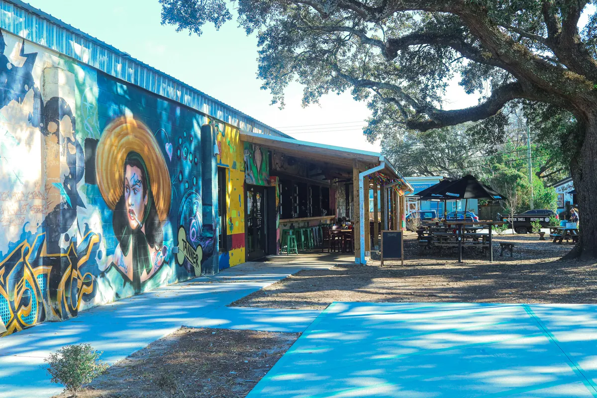 A colorful mural adjacent to an outdoor seating area beneath a large oak tree, with people dining and gathering. The scene exudes an artistic, energetic vibe characteristic of local eateries and social spaces.