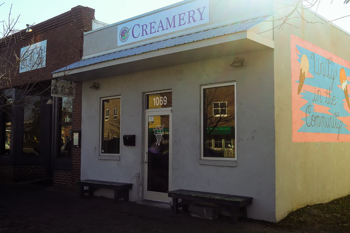 The image features the exterior of a local creamery, highlighted by a cheerful community mural and inviting benches near the entrance. The area is lively and approachable, suggesting a popular neighborhood hangout spot perfect for socializing with friends or enjoying a treat after an evening out.