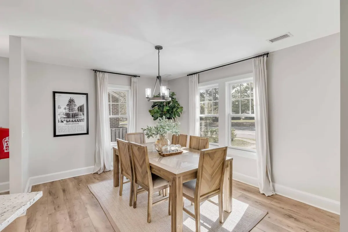 A stylish dining area featuring a large dining table, elegant chandelier, expansive windows letting in natural light, a plant centerpiece, and wall art. The setting is arranged for group meals and social gatherings, radiating warmth and hospitality.